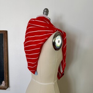 May include: A bright red scarf featuring thin horizontal white stripes draped over a beige dress form. The fabric is lightweight and soft, showing a consistent pattern of stripes across the entire surface of the rectangular textile accessory.