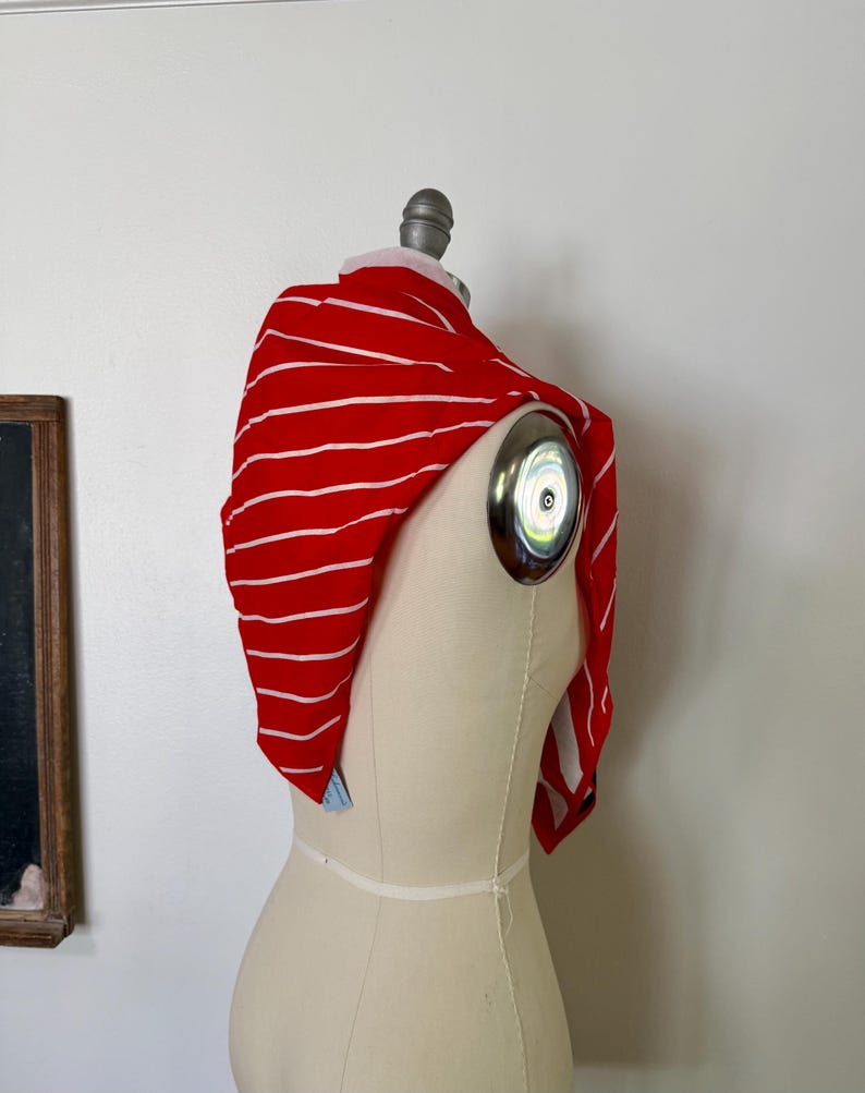 May include: A bright red scarf featuring thin horizontal white stripes draped over a beige dress form. The fabric is lightweight and soft, showing a consistent pattern of stripes across the entire surface of the rectangular textile accessory.
