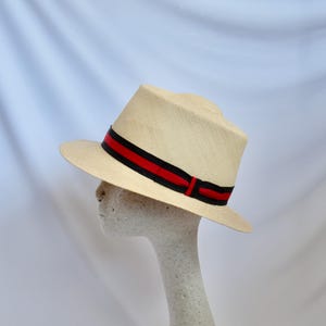 May include: A light-colored fedora hat with a black and red striped band. The hat is displayed on a mannequin head against a white background. The hat's brim is slightly upturned.