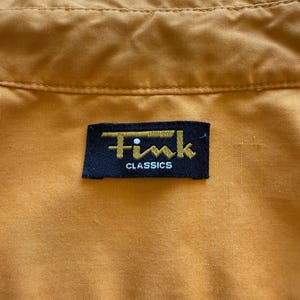 May include: Close-up of a mustard-colored garment with a black label. The label reads "Fink CLASSICS" in gold lettering. The fabric has a subtle texture and visible stitching.