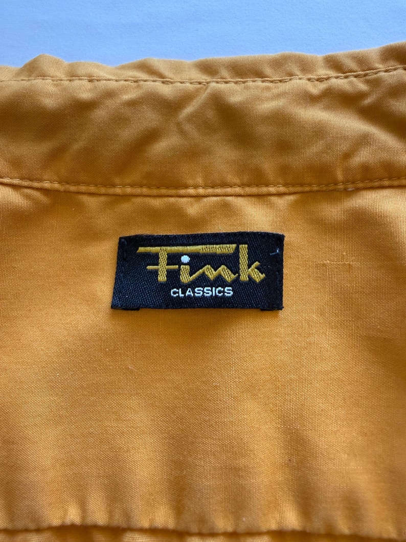May include: Close-up of a mustard-colored garment with a black label. The label reads "Fink CLASSICS" in gold lettering. The fabric has a subtle texture and visible stitching.