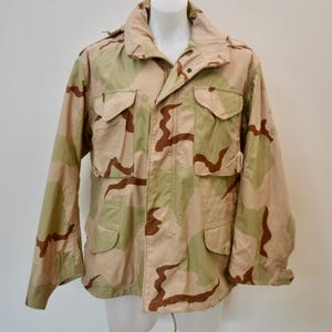 Könnte beinhalten: Eine Feldjacke mit Wüstentarnmuster in den Farben Beige, Hellbraun und Olivgrün. Die Jacke verfügt über einen Umlegekragen, einen Frontreißverschluss mit Druckknopfleiste sowie vier große aufgesetzte Taschen mit geknöpften Patten auf der Vorderseite.
