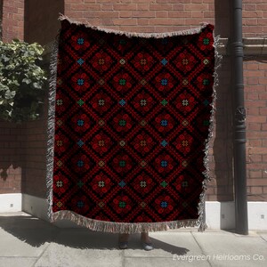 A person is standing in front of a brick wall, holding a large, colorful, patterned blanket with a red and black design.