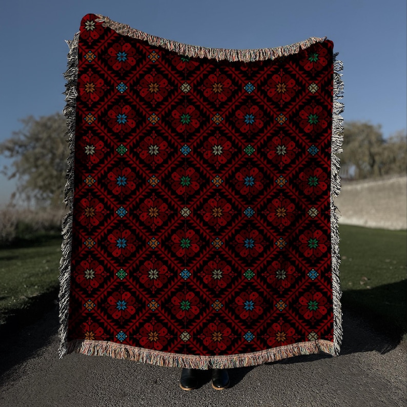 A person is standing in front of a large, colorful, patterned blanket with a red and black design.