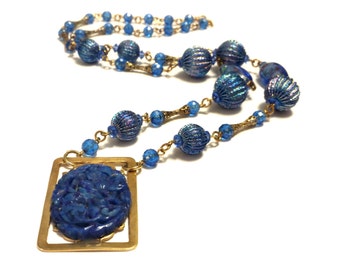 Puede incluir: Un collar azul y dorado con un colgante de piedra azul tallada. El collar presenta una cadena con cuentas de vidrio azul y cuentas azules más grandes con una superficie texturizada.