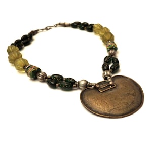 May include: A necklace with a heart-shaped pendant made of brass with intricate engravings. The necklace is made of green and yellow glass beads and silver spacers.