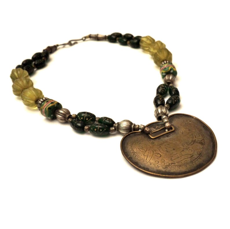 May include: A necklace with a heart-shaped pendant made of brass with intricate engravings. The necklace is made of green and yellow glass beads and silver spacers.