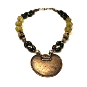 May include: A silver necklace with a large, heart-shaped pendant. The pendant is made of brass and has an intricate design. The necklace is strung with green and yellow glass beads.