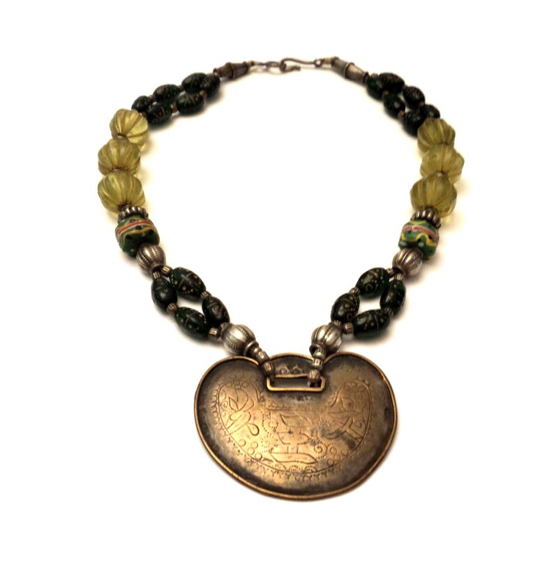 May include: A silver necklace with a large, heart-shaped pendant. The pendant is made of brass and has an intricate design. The necklace is strung with green and yellow glass beads.