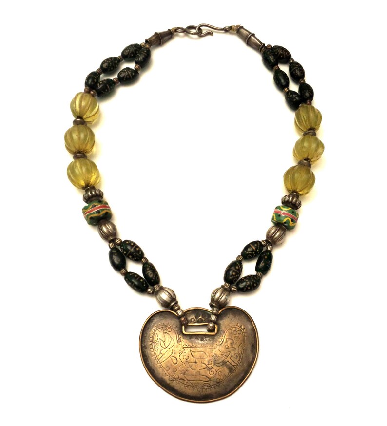 May include: A silver necklace with a heart-shaped pendant. The pendant is decorated with intricate engravings. The necklace is made with green and yellow glass beads.