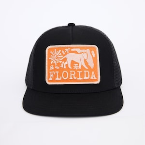 May include: Black trucker hat with a large orange patch featuring a white silhouette of a Florida panther and the word "FLORIDA" in white text.