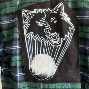May include: A green, blue, and black plaid flannel shirt. A black patch on the back features a white outline of a wolf's head with a white sphere and radiating lines. The shirt is made of a soft, comfortable material.