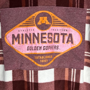 May include: A maroon and pink graphic featuring the text "MINNESOTA GOLDEN GOPHERS" with the university's logo. The graphic is on a maroon fabric, set against a plaid background. The words "ATHLETIC TRADITION" and "ESTABLISHED 1851" are also visible.