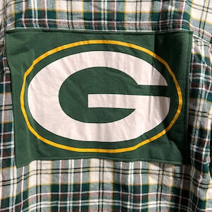 May include: A long-sleeve button-down shirt featuring a green, white, and yellow plaid pattern. A large, rectangular green fabric patch is sewn onto the back, displaying a prominent white letter G logo outlined in yellow and green.