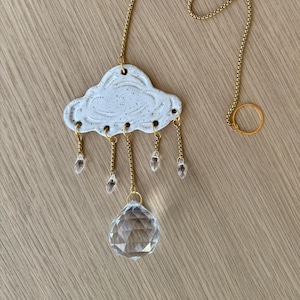 May include: A white ceramic cloud-shaped hanging decoration with gold-colored chain and crystal accents. The cloud has a speckled texture and hangs from a gold chain with a ring at the top. Clear crystal droplets hang from the cloud.