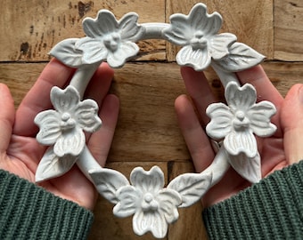 May include: A circular ceramic trivet featuring a speckled off-white glaze with four sculpted dogwood flowers evenly spaced around the ring. Each flower has four petals and a central cluster of three small buds, accompanied by sculpted leaves.