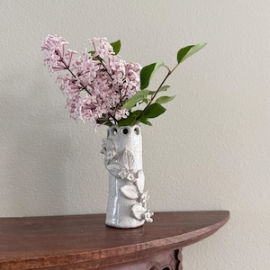 May include: A cylindrical ceramic vase with a speckled white glaze, featuring a decorative relief of sculpted leaves and small flowers on the exterior. The top rim has a series of circular cutouts, and it holds a cluster of light purple lilac flowers.