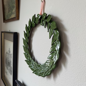 Pode incluir: Um adorno circular de parede composto por uma coroa de folhas de cerâmica em verde-escuro brilhante. A coroa está suspensa por uma fita de veludo rosa-claro, fixada com um pequeno rebite de metal prateado.