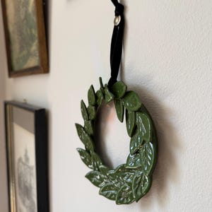 Small green ceramic leaf wreath hanging on a wall beside framed vintage art, styled as gallery wall decor.