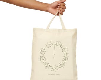 May include: A cream-colored tote bag with a floral wreath design in black ink. The bag is held by a hand and features the text "Julia Helene Pottery" at the bottom. The tote bag has two handles and is made of a sturdy fabric.