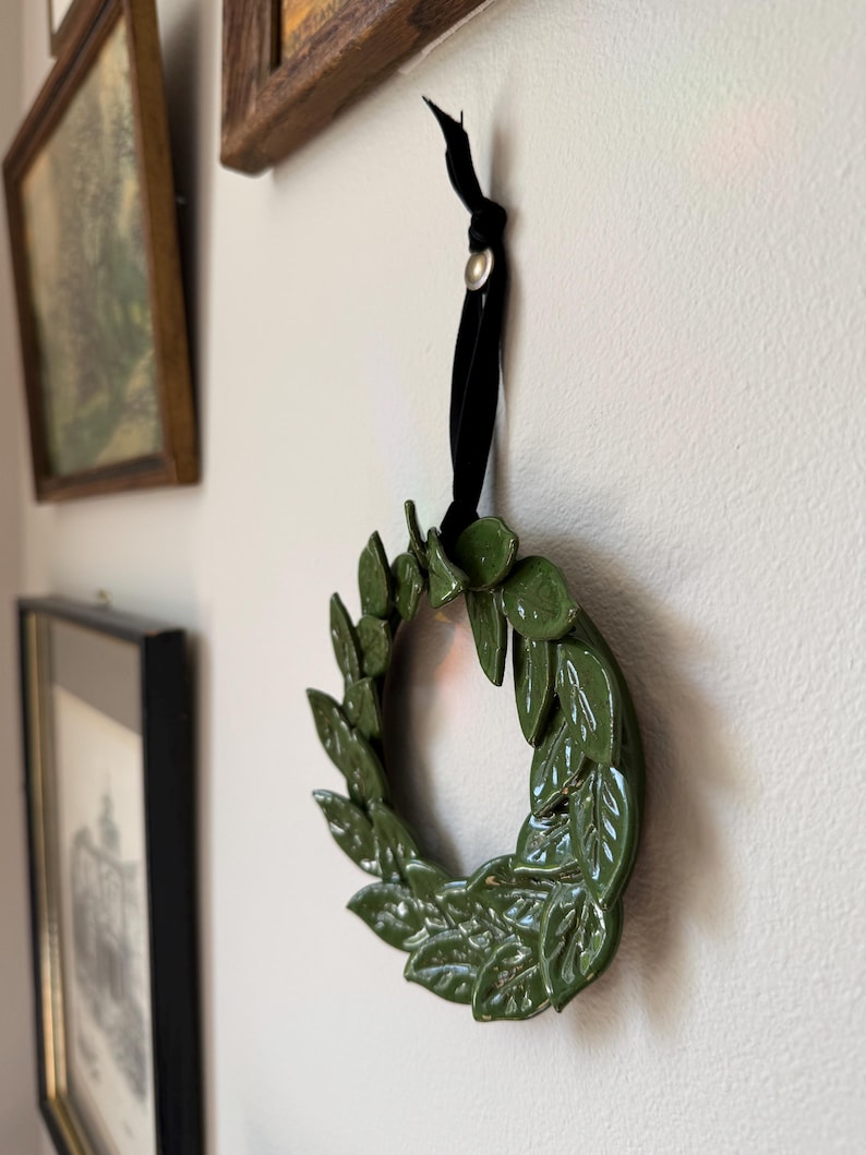 Small green ceramic leaf wreath hanging on a wall beside framed vintage art, styled as gallery wall decor.