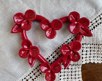 May include: A glossy red heart-shaped wall hanging crafted from a smooth material. The heart frame is decorated with five distinct three-petaled flower clusters, each featuring small pointed leaves attached to the curved red structure.