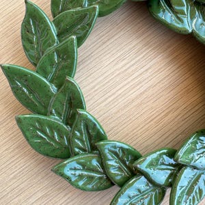 A circular wreath composed of individual ceramic leaves finished in a glossy, clover green glaze. Each leaf features visible veining and subtle dark brown speckling throughout the surface, arranged in an overlapping, layered pattern.