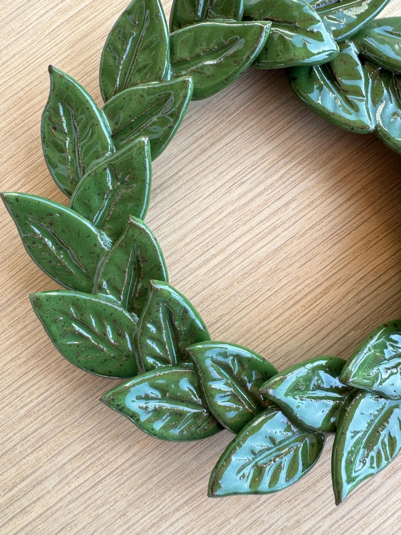 A circular wreath composed of individual ceramic leaves finished in a glossy, clover green glaze. Each leaf features visible veining and subtle dark brown speckling throughout the surface, arranged in an overlapping, layered pattern.