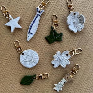 May include: A collection of eight ceramic charms with gold-toned lobster clasps. The shapes include a white speckled star, a blue and white fish, a dark green ivy leaf, a white flower, a round white speckled disc, a white flower, a green leaf, and a star.