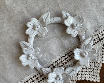 May include: A white ceramic floral wreath with speckled detailing. The wreath features five dimensional flowers and leaves. The wreath is placed on a beige linen surface with a lace trim.