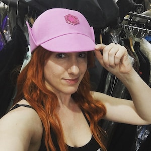 May include: A pink baseball cap with a pink and white symbol on the front. The cap has a short brim and a pointed ear flap on each side.