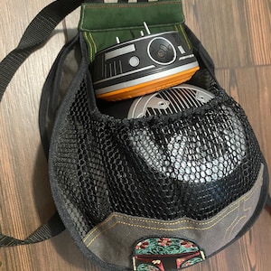 May include: A black mesh backpack with a green fabric flap and a brown leather strap. The backpack has a small, round, black and orange toy robot inside. The backpack also has a small embroidered patch of a Star Wars character.