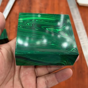 May include: A square malachite stone with a polished surface. The stone is a vibrant green with swirling patterns of lighter and darker shades, creating a natural, marbled effect. The stone is held in a hand.
