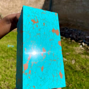 Puede incluir: Una piedra rectangular turquesa con vetas e inclusiones marrones. La piedra, sostenida en una mano, tiene una superficie pulida. La piedra es de un color azul-verdoso vibrante y es probablemente un objeto decorativo o una materia prima.