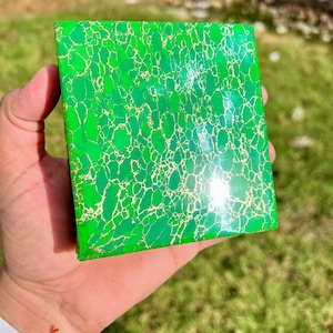 May include: A square, green box with a gold, cracked-pattern design. The box is held in a person's hand against a background of green grass. The box has a glossy finish and appears to be made of a solid material.