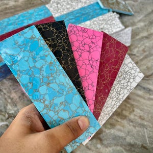 Puede incluir: Una mano sostiene un abanico de baldosas de piedra decorativas rectangulares. Las baldosas muestran una variedad de colores, incluyendo turquesa, negro, rosa y burdeos, cada una con un diseño único. Las baldosas son probablemente para manualidades o fines decorativos.