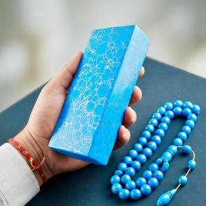 Puede incluir: Un objeto azul de forma geométrica con un patrón blanco similar a una telaraña se sostiene en una mano. Un collar de cuentas de oración azules con una borla decorativa está cerca. Las cuentas son de un tono azul similar.