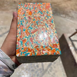 Puede incluir: Un bloque rectangular con una superficie de mosaico compuesta por pequeñas piezas angulares de color naranja, blanco y turquesa, intercaladas con finas escamas metálicas de color dorado reflectante en un acabado liso y brillante.
