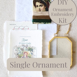 May include: A DIY ornament embroidery kit featuring a printed card with the text Anne of Green Gables, a white felt square, embroidery floss in beige, brown, and green, and a gold-toned metal frame with a chain for hanging a single ornament.