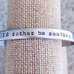May include: A silver-colored cuff bracelet with the stamped phrase "i'd rather be reading." The bracelet is displayed on a beige, textured surface. The text is in a simple, sans-serif font.
