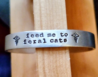Może przedstawiać: Srebrna bransoletka z napisem "feed me to feral cats" i dwoma symbolami medycznymi po obu stronach.