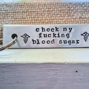 May include: A rectangular metal keychain with the stamped text "check my fucking blood sugar". The keychain has a key ring attached and medical symbols on each side of the text.