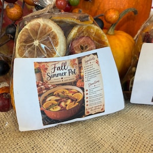 Puede incluir: Una bolsa de plástico transparente con rodajas de naranja y trozos de manzana secos, atada con cuerda. Una etiqueta blanca muestra el texto Fall Simmer Pot, Warm Scents, Cozy Spaces, junto con una lista de ingredientes e instrucciones.