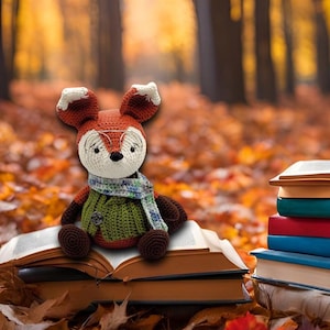 May include: A crocheted fox wearing glasses and a scarf sits on a stack of books in a forest setting with autumn leaves on the ground.