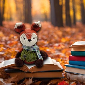 May include: A crocheted fox wearing glasses and a scarf sits on a stack of books in a forest setting with autumn leaves on the ground.