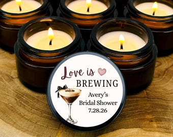 Peut inclure: Sept bougies allumées dans de petits pots en verre brun sont disposées sur une surface en bois. L'une des bougies porte une étiquette avec le texte "Love is Brewing", une illustration de cocktail et "Avery's Bridal Shower 7.28.26".