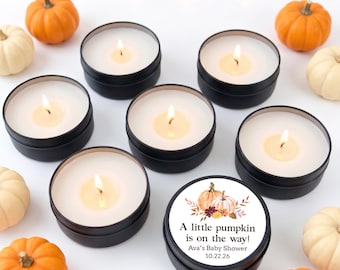 Op de afbeelding: Zes zwarte metalen blikjes met witte kaarsen met brandende lonten, omringd door kleine oranje en witte pompoenen. Eén deksel heeft een label met een pompoenillustratie en de tekst: A little pumpkin is on the way! Ava's Baby Shower 10.22.26.