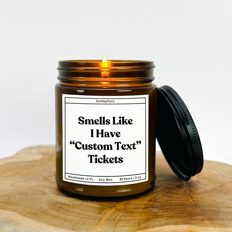 May include: A brown glass candle jar with a black lid. The candle is lit and the label reads "Smells Like I Have "Custom Text" Tickets". The label also includes "Handmade in FL, Soy Wax, 40 hours/9 oz".