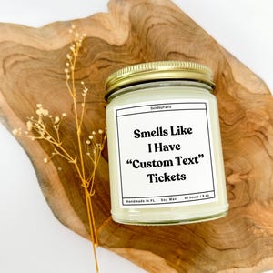 May include: A white candle with a gold lid and a white label that reads "Sunday Faire" and "Smells Like I Have "Custom Text" Tickets". The label also includes "Handmade in FL", "Soy Wax", and "40 hours/9 oz".
