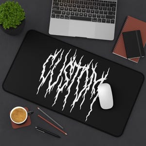 May include: Black rectangular desk mat with a white abstract design. A white computer mouse sits on the mat, near a laptop. A small potted plant, notebooks, and a cup of coffee are also visible.